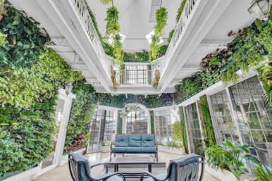 Bright conservatory-style sunroom with floor-to-ceiling living green walls, hanging vines and skylight, white balcony railings, and blue cushioned sofa and chairs around a coffee table.