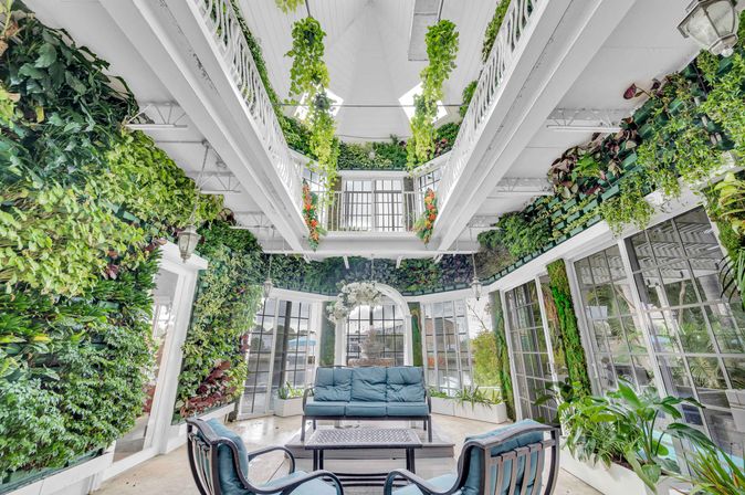 Bright conservatory-style sunroom with floor-to-ceiling living green walls, hanging vines and skylight, white balcony railings, and blue cushioned sofa and chairs around a coffee table.