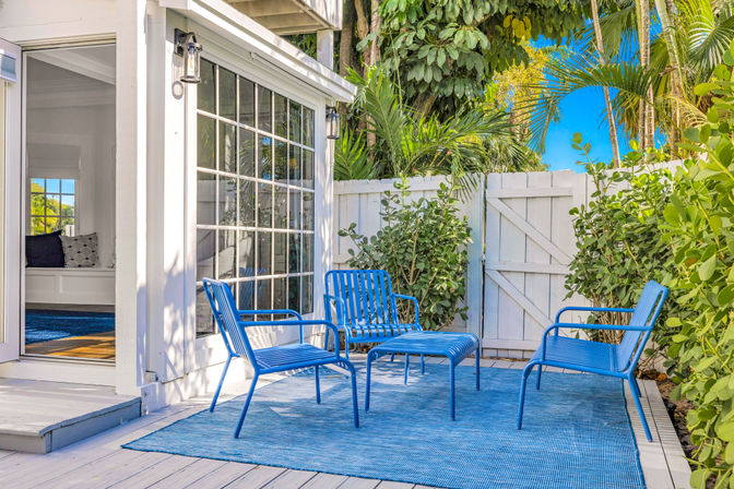Sunny tropical backyard patio with bright blue metal chairs and matching ottoman on a blue rug over white wood decking, beside large grid-pane sliding glass doors, a white picket-style gate, and lush palm and hedge foliage under a clear blue sky.