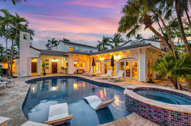 Tropical luxury villa at sunset with illuminated curved swimming pool and attached spa, poolside loungers and umbrellas, palm trees and tiled patio