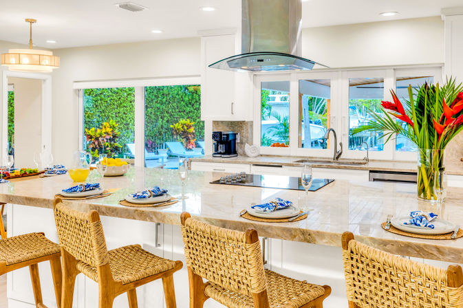 Bright coastal open-concept kitchen with large marble island, woven rattan bar stools, set place settings, stainless range hood and sliding glass doors overlooking pool and tropical landscaping