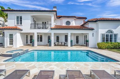 Sunny Mediterranean-style white two-story villa with red tile roof, backyard swimming pool and spa, tiled patio with lounge chairs and outdoor dining, wrought-iron balcony and blue sky