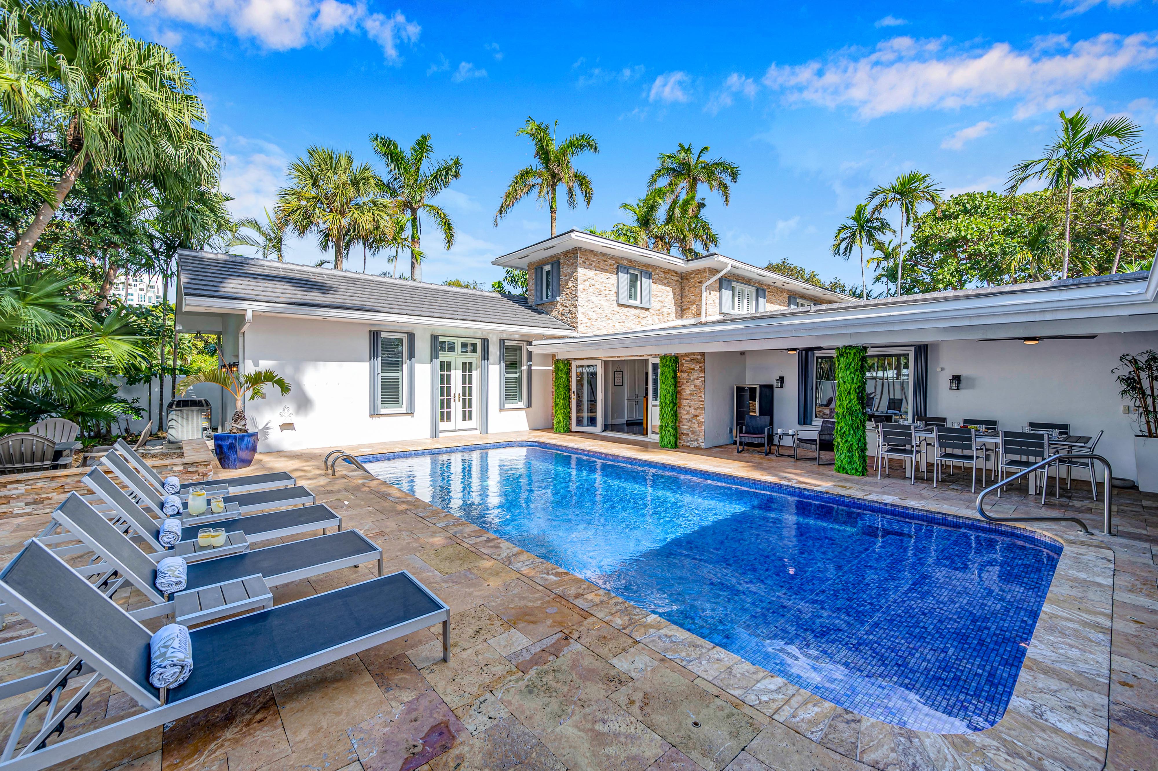 Sunny tropical backyard oasis with a rectangular blue-tiled swimming pool, row of lounge chairs with rolled towels on a travertine patio, palm trees, and a modern two-story white pool house with covered dining and seating area.