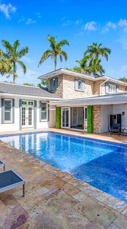 Sunny tropical backyard oasis with a rectangular blue-tiled swimming pool, row of lounge chairs with rolled towels on a travertine patio, palm trees, and a modern two-story white pool house with covered dining and seating area.