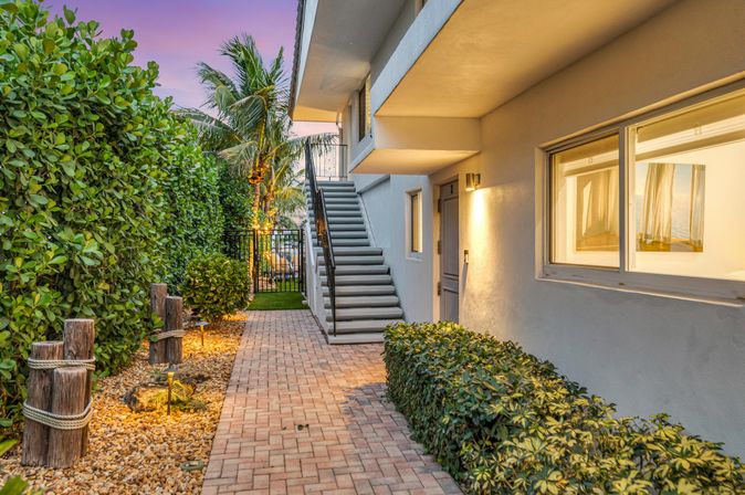 Brick walkway leading to an exterior staircase in a coastal apartment courtyard at sunset, with lit tropical landscaping, palms and trimmed hedges.