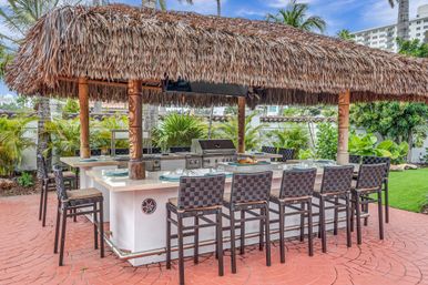 Tropical outdoor tiki bar and kitchen with a thatched palapa, built-in stainless grill and island counter lined with woven bar stools on a red brick patio, surrounded by palm trees and lush greenery.