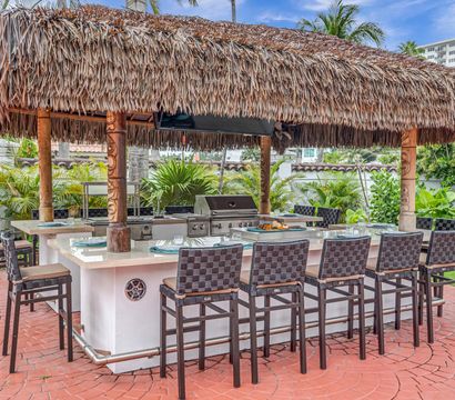 Tropical outdoor tiki bar and kitchen with a thatched palapa, built-in stainless grill and island counter lined with woven bar stools on a red brick patio, surrounded by palm trees and lush greenery.