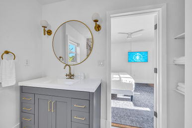 Bright modern bathroom with gray vanity, white countertop, round gold-framed mirror and brass fixtures; open door reveals a sunlit ensuite bedroom with wall-mounted TV and ceiling fan.