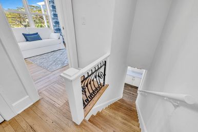 Bright upstairs landing with hardwood stairs and white railing featuring decorative black iron spindles, opening to a sunlit room with a white sofa, blue pillows, patterned rug and large windows.