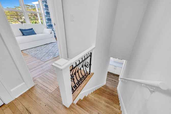 Bright upstairs landing with hardwood stairs and white railing featuring decorative black iron spindles, opening to a sunlit room with a white sofa, blue pillows, patterned rug and large windows.