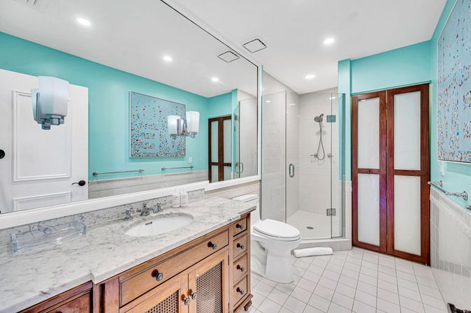 Cheerful turquoise bathroom with marble countertop and undermount sink, wooden vanity, frameless glass shower with handheld showerhead, white toilet, large wall mirror and colorful abstract artwork.