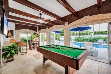 Sunlit poolside game room with green billiards table under wood-beamed ceiling and stone arches, open glass doors to tiled patio with lounge chairs, blue umbrellas and a swimming pool