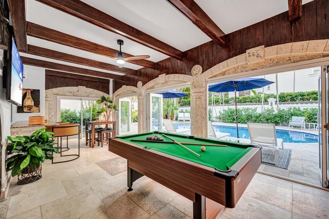 Sunlit poolside game room with green billiards table under wood-beamed ceiling and stone arches, open glass doors to tiled patio with lounge chairs, blue umbrellas and a swimming pool