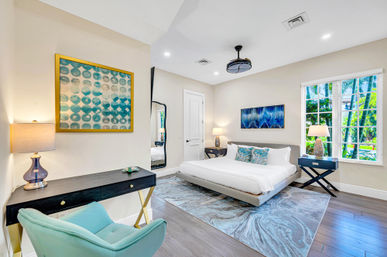 Bright modern coastal bedroom with low platform king bed, turquoise accent pillows, abstract wall art, black desk and bedside tables, teal armchair, marbled area rug, hardwood floors and large window looking out at palm trees
