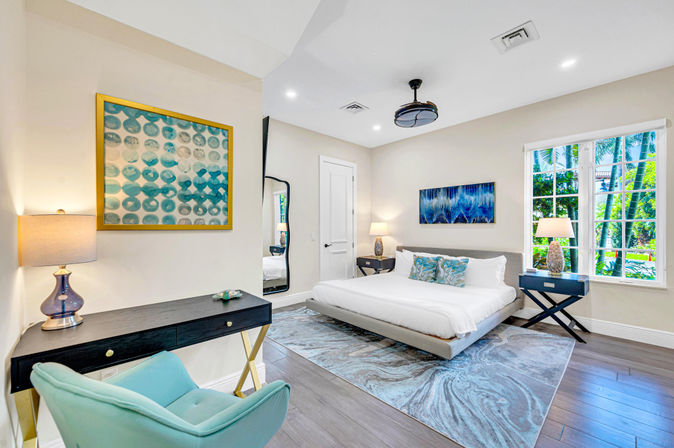 Bright modern coastal bedroom with low platform king bed, turquoise accent pillows, abstract wall art, black desk and bedside tables, teal armchair, marbled area rug, hardwood floors and large window looking out at palm trees