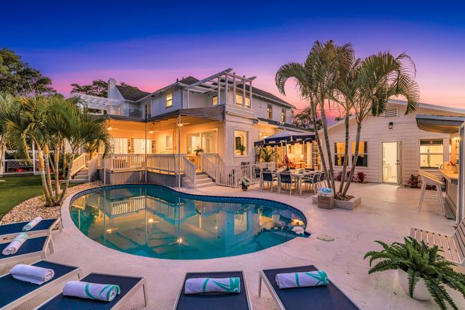 Twilight view of a tropical coastal vacation-home backyard with a lit two-story house, palm trees, kidney-shaped pool, lounge chairs with rolled towels, and outdoor dining under a purple-pink sunset.