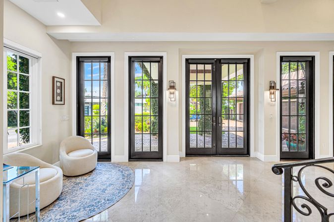 Bright luxury foyer with black-framed French doors and sidelights opening to a tropical garden, glossy marble floor, two cream swivel chairs on a blue patterned rug, and a wrought-iron staircase railing.