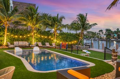 Sunset waterfront resort-style backyard pool framed by palm trees and glowing string lights, with lounge chairs and a marina dock with boats