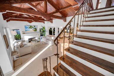 Sunlit Mediterranean-style living room with dark exposed wooden beams, marble floors, cream sofas, fireplace and wrought-iron staircase in a luxury coastal interior.