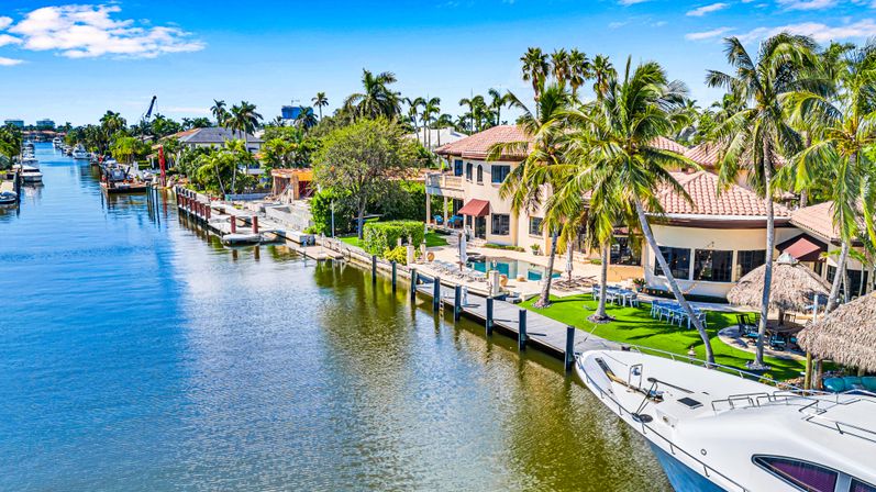 White yacht bow in foreground on a sunny Florida waterfront canal lined with palm trees, private docks and Mediterranean-style luxury homes under a bright blue sky