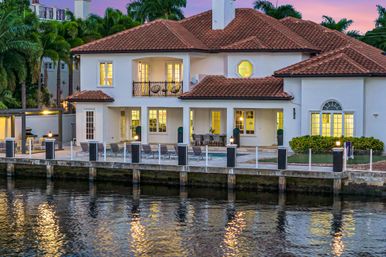 Mediterranean-style canalfront house with red tile roof, glowing windows, pool and patio with lounge chairs, private dock and palm trees at sunset.