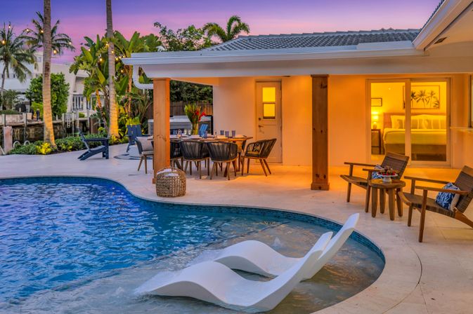 Tropical waterfront backyard at dusk with curved pool and in-water loungers, covered outdoor dining area, warm interior glow, palm trees and canal