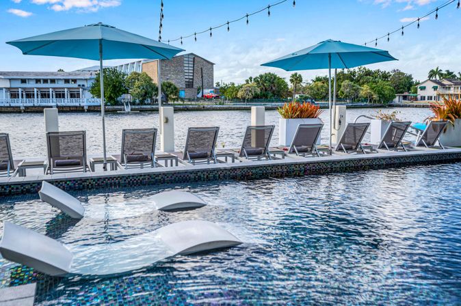 Waterfront pool deck with turquoise umbrellas, a row of lounge chairs and planters along a canal, in-pool sunbeds and string lights overhead