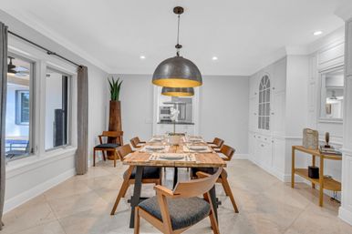 Modern dining room interior with a long wooden table set for six, mid-century wooden chairs, oversized black-and-gold pendant lights, white built-in cabinetry, beige tile floor, and large windows with gray curtains.