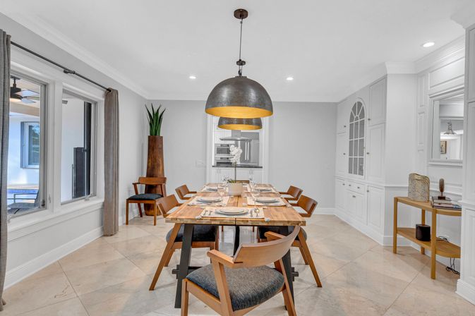 Modern dining room interior with a long wooden table set for six, mid-century wooden chairs, oversized black-and-gold pendant lights, white built-in cabinetry, beige tile floor, and large windows with gray curtains.