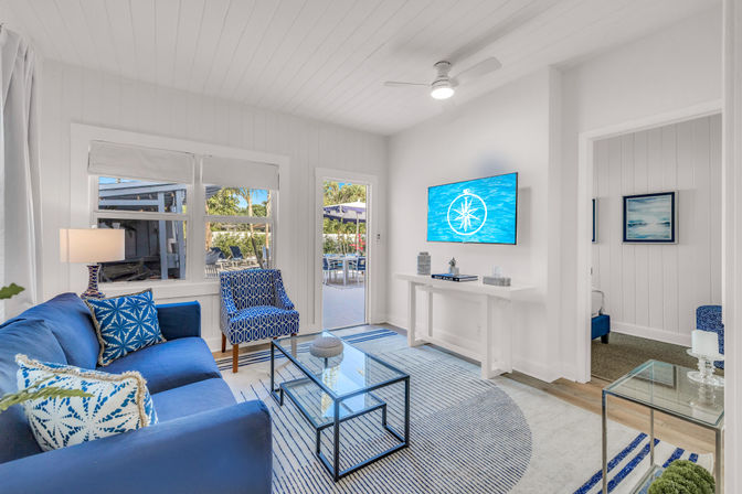 Bright coastal living room with white shiplap walls, blue sofa and patterned armchair, glass coffee table on a striped round rug, wall-mounted TV with nautical compass graphic, and a door opening to a sunny patio dining area.