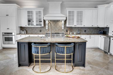 Bright modern white kitchen with granite island, navy velvet bar stools with gold frames, stainless appliances, decorative range hood and diamond-tile backsplash.