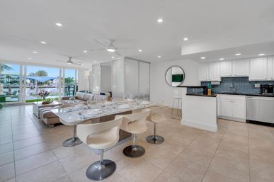 Open-plan modern waterfront condo interior with dining table set, white kitchen, leather sectional and marina view through floor-to-ceiling sliding glass doors.