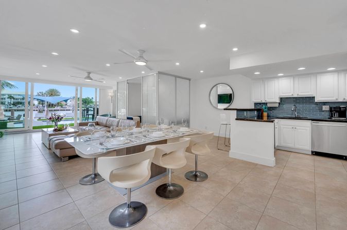 Open-plan modern waterfront condo interior with dining table set, white kitchen, leather sectional and marina view through floor-to-ceiling sliding glass doors.