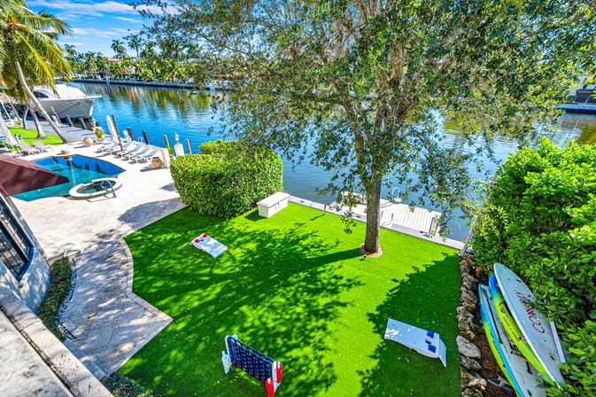 Sunlit waterfront backyard on a calm canal with a pool and spa, sun loungers and dock, palm trees, grassy play area with cornhole and giant Connect Four, and stacked paddleboards.