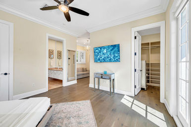 Sunlit master bedroom with hardwood floors, modern ceiling fan, wall-mounted TV above a mirrored console, open en-suite bathroom and walk-in closet, and French doors letting in natural light.