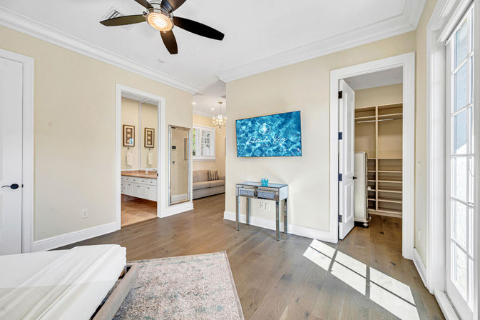 Sunlit master bedroom with hardwood floors, modern ceiling fan, wall-mounted TV above a mirrored console, open en-suite bathroom and walk-in closet, and French doors letting in natural light.