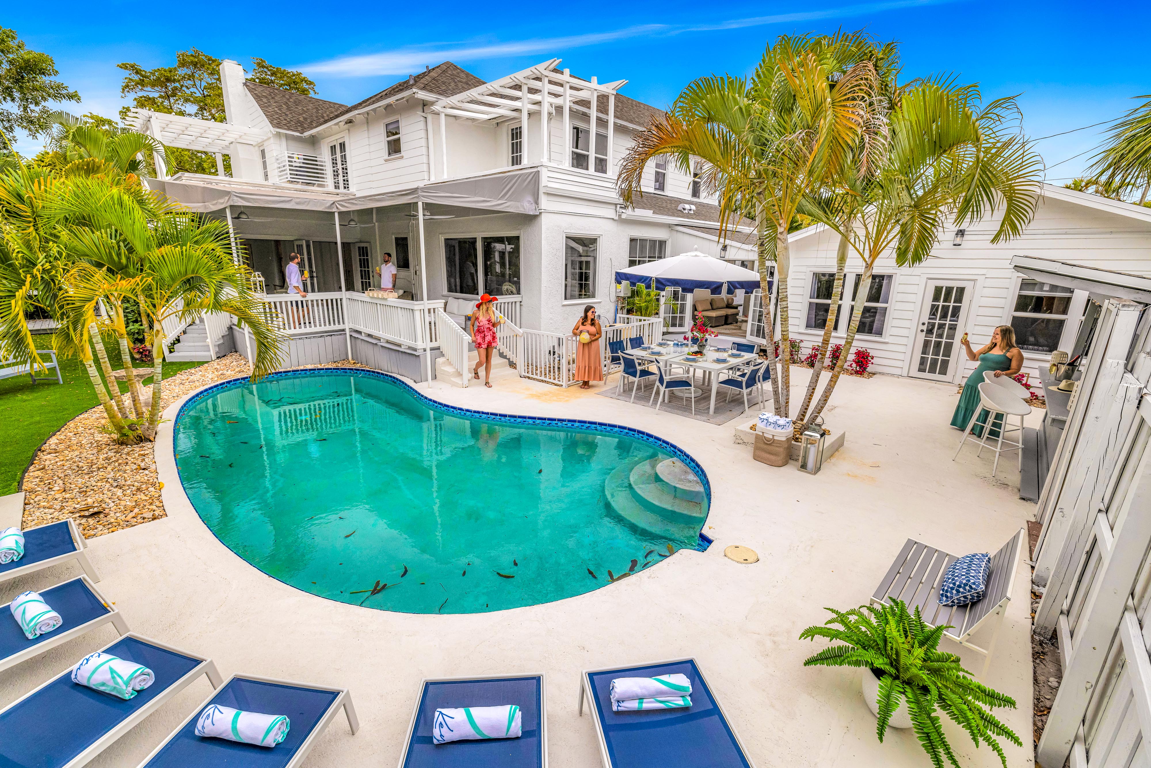 Sunny Florida-style backyard with kidney-shaped turquoise pool, white coastal house, palm trees, lounge chairs and poolside dining with people