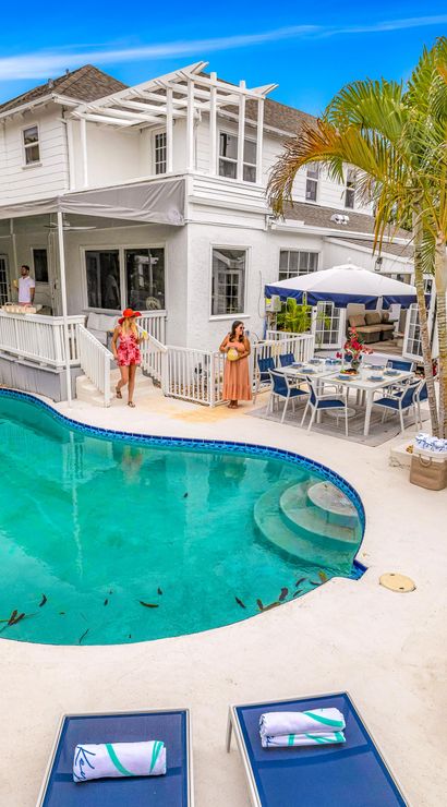 Sunny Florida-style backyard with kidney-shaped turquoise pool, white coastal house, palm trees, lounge chairs and poolside dining with people