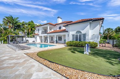 Luxury Mediterranean-style two-story home with red tile roof, backyard swimming pool, row of loungers and cabana, small putting green, palm trees and bright blue sky