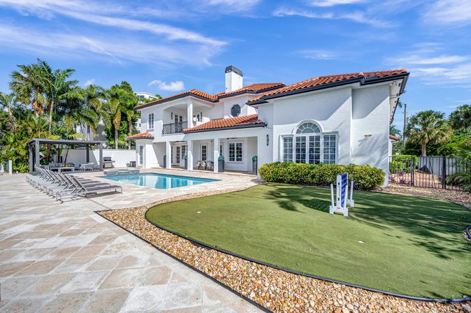 Luxury Mediterranean-style two-story home with red tile roof, backyard swimming pool, row of loungers and cabana, small putting green, palm trees and bright blue sky