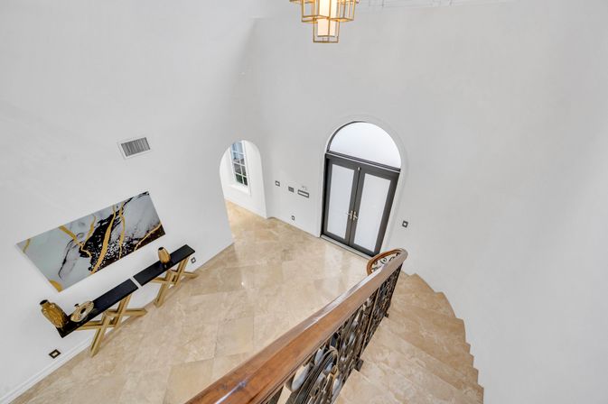 Overhead view of a sunlit luxury foyer with polished marble floors, arched black double doors, curved staircase with ornate wrought-iron railing, black console table with gold accents beneath abstract wall art, and a geometric chandelier.