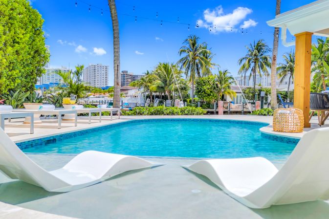 Tropical waterfront pool with curved turquoise water, submerged white lounge chairs, palm trees, string lights and low‑rise city buildings under a bright blue sky