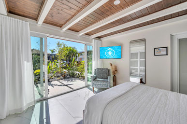 Sunlit coastal bedroom with wood-beamed ceiling, floor-to-ceiling sliding glass doors opening to a tropical courtyard garden, patterned accent chair and wall-mounted TV