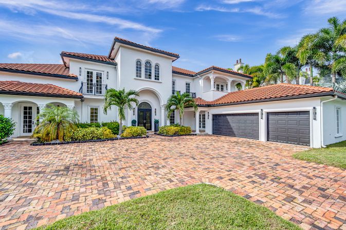 Sunlit Mediterranean-style luxury home with terracotta tile roof, arched entry, palm trees and three-car garage on a brick paver driveway in a tropical setting