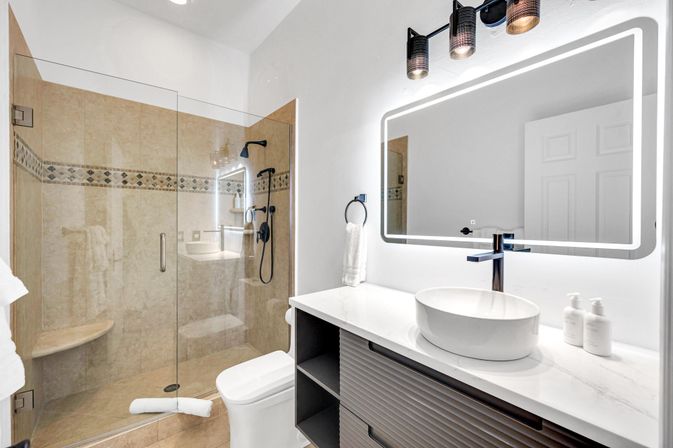 Modern spa-like master bathroom with glass walk-in shower, beige tiled walls with decorative border, matte black fixtures, LED-lit vanity mirror, white vessel sink on a sleek white countertop.