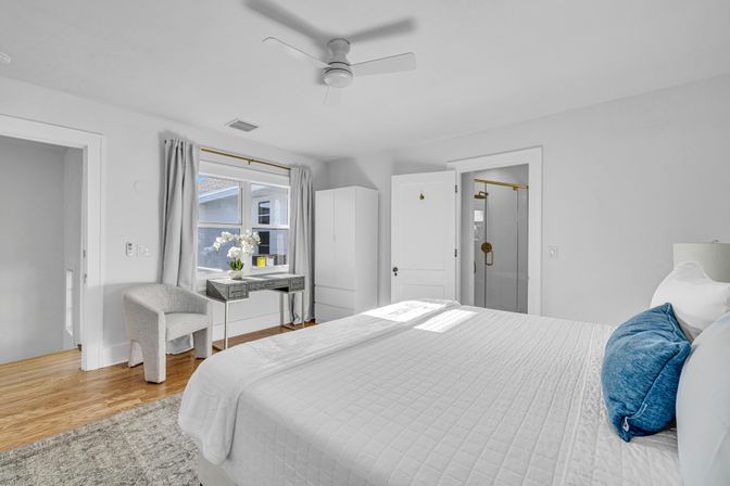 Sunlit modern master bedroom with white quilted bed and blue accent pillows, hardwood floors, ceiling fan, desk with orchid by the window, gray armchair, wardrobe, and en-suite glass shower with brass fixtures.