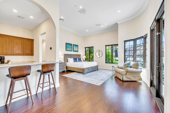 Bright open-plan master bedroom with hardwood floors, king bed and nightstands, kitchen counter with two wooden bar stools, cozy armchair by bay windows and French doors overlooking a lush tropical patio — modern coastal style with abundant natural light.