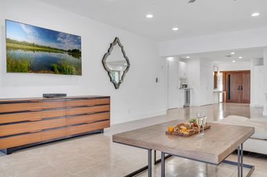 Bright modern open-plan living room with white walls and tile floors, wall-mounted TV over a mid-century wood dresser, decorative mirror, wooden coffee table with charcuterie board and two champagne flutes, and wood entry doors in the background.