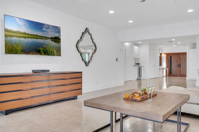Bright modern open-plan living room with white walls and tile floors, wall-mounted TV over a mid-century wood dresser, decorative mirror, wooden coffee table with charcuterie board and two champagne flutes, and wood entry doors in the background.