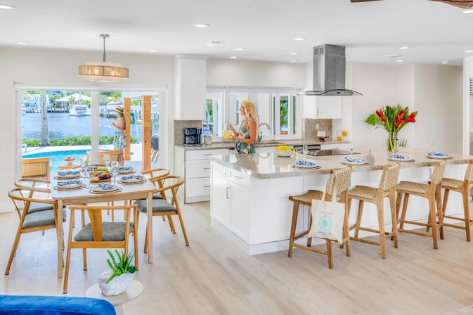 Sunlit coastal open-plan kitchen and dining area with a marble island, wicker bar stools, table set for a meal, sliding doors to a pool and waterfront view, and a person pouring juice at the sink.